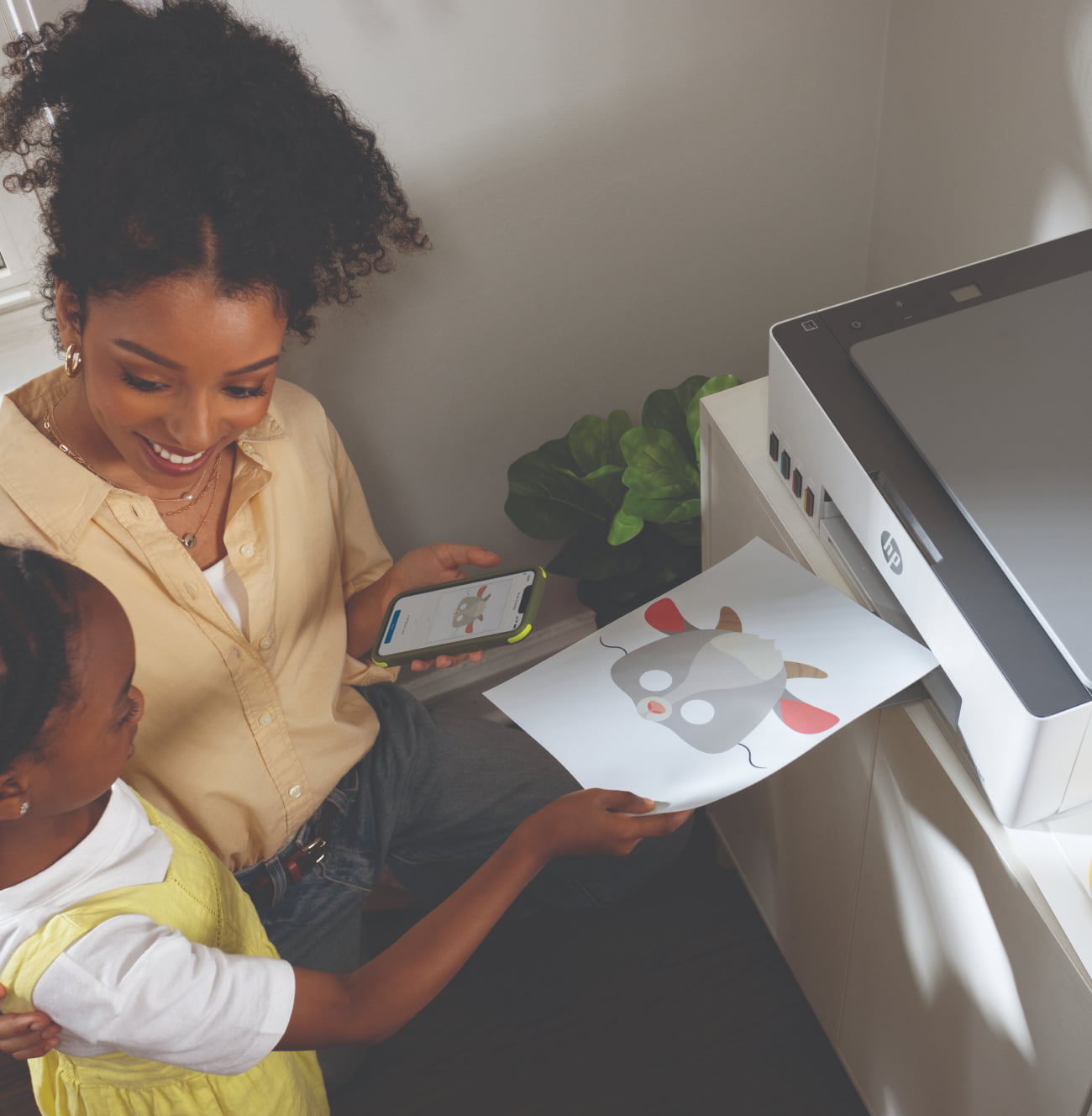 Mother with child sitting near the HP Smart Tank printer and printing colorful pictures using the app.